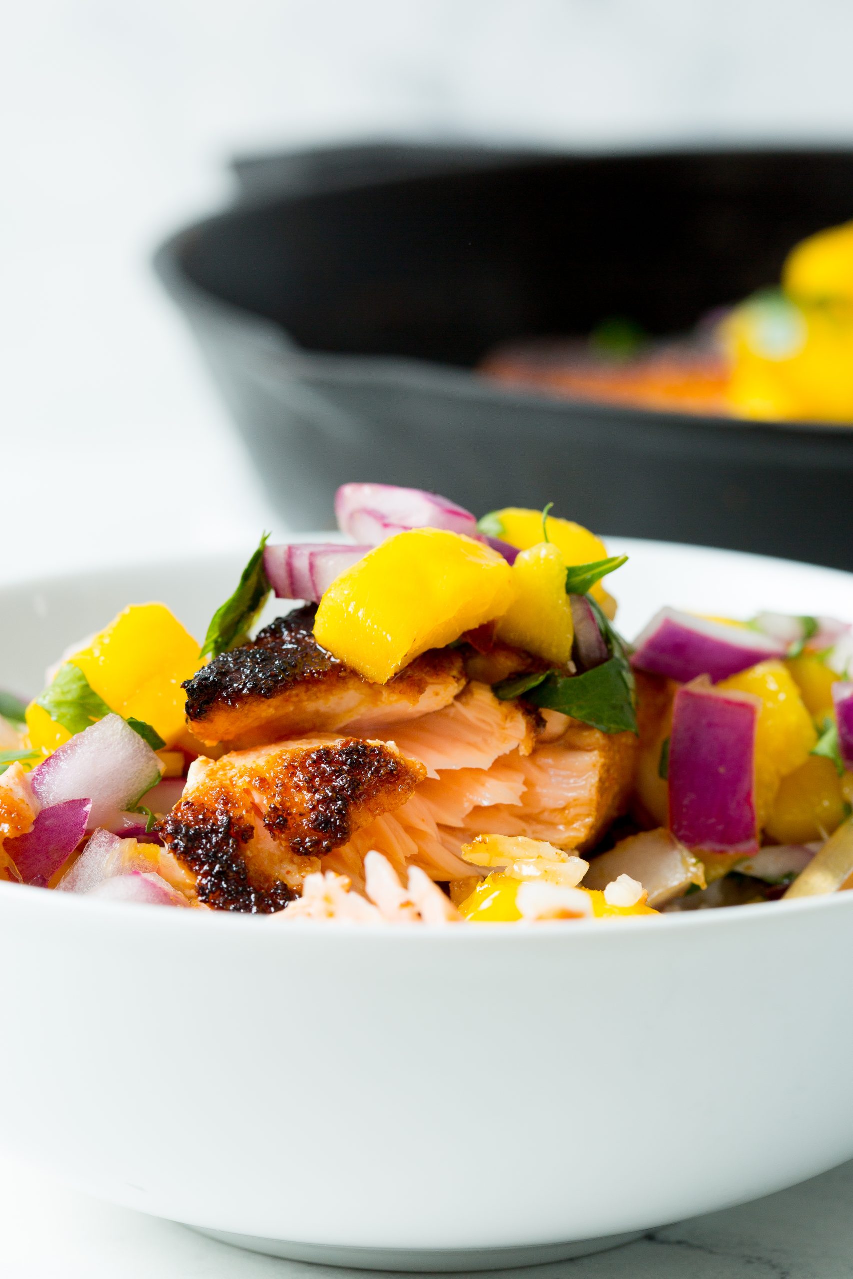 Bowl of grilled fish topped with diced fruit and herbs, with a skillet in the background.