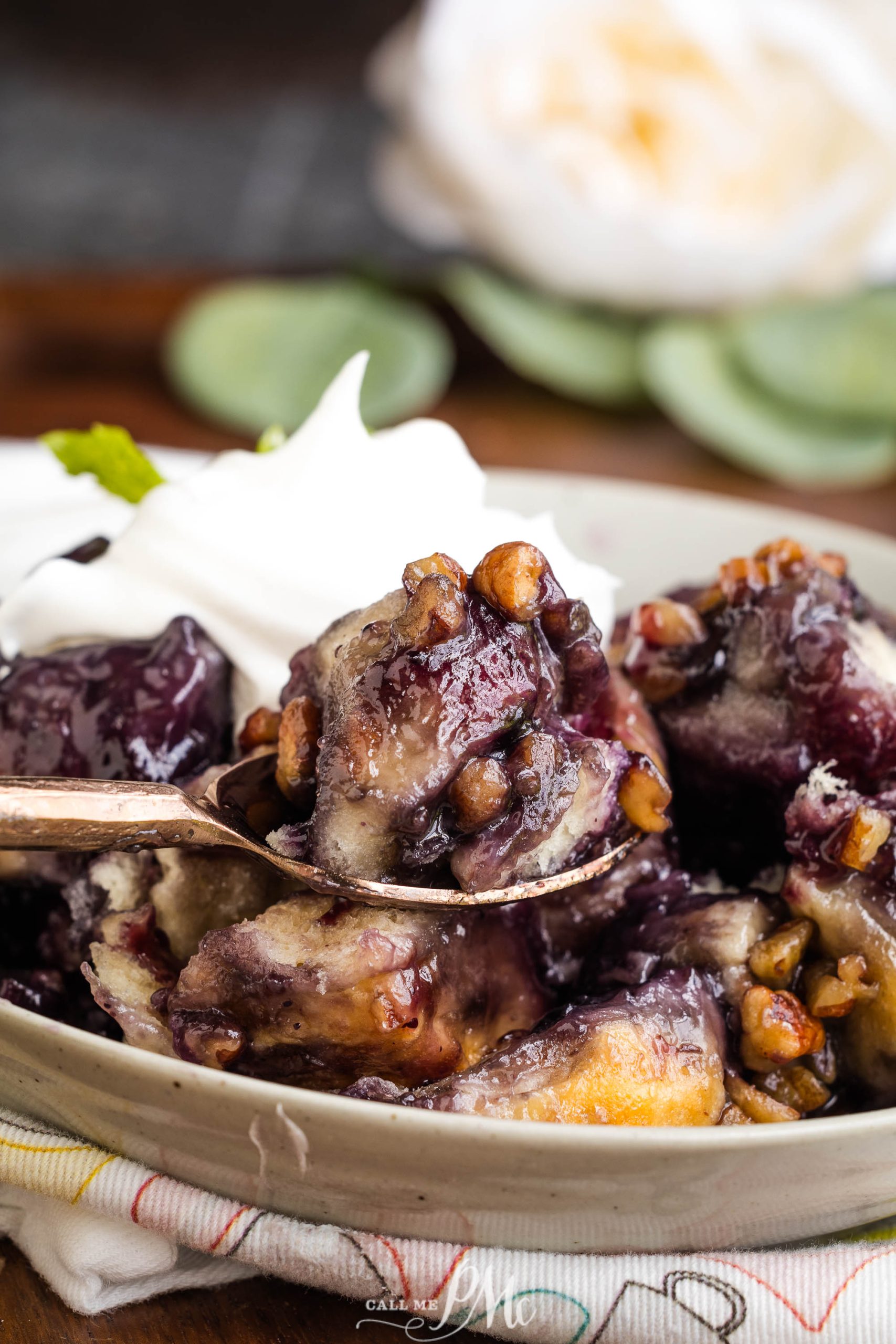 A close-up of blueberry cobbler in a bowl, topped with pecans and whipped cream. A spoon is lifting a portion of the dessert.