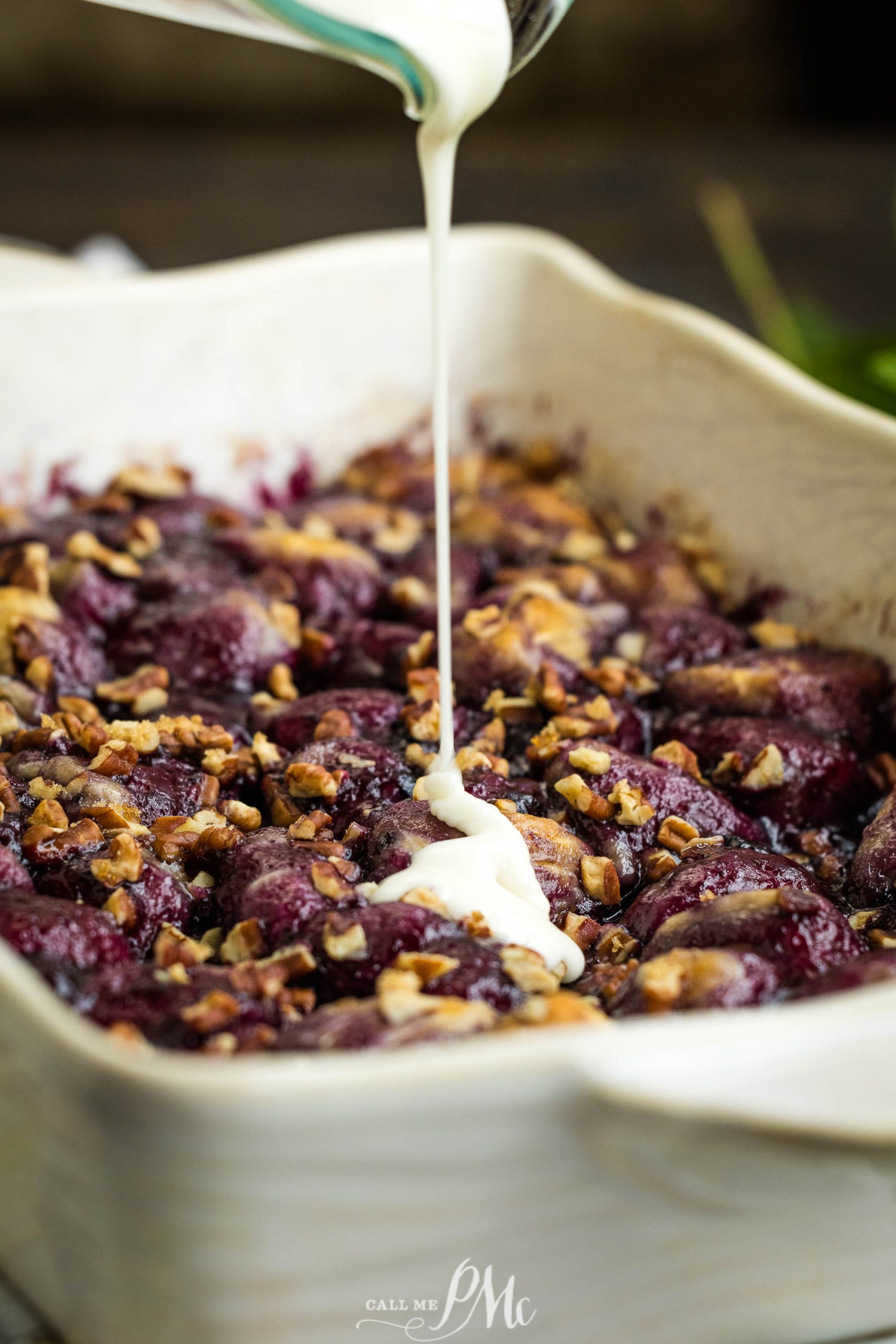 A baking dish filled with a berry and nut mixture is being drizzled with a white cream sauce.