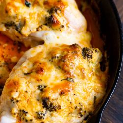 Close-up of two baked chicken breasts in a cast iron skillet, topped with melted cheese and herbs. The dish has a golden-brown crust with visible spices and is sitting on a wooden surface.