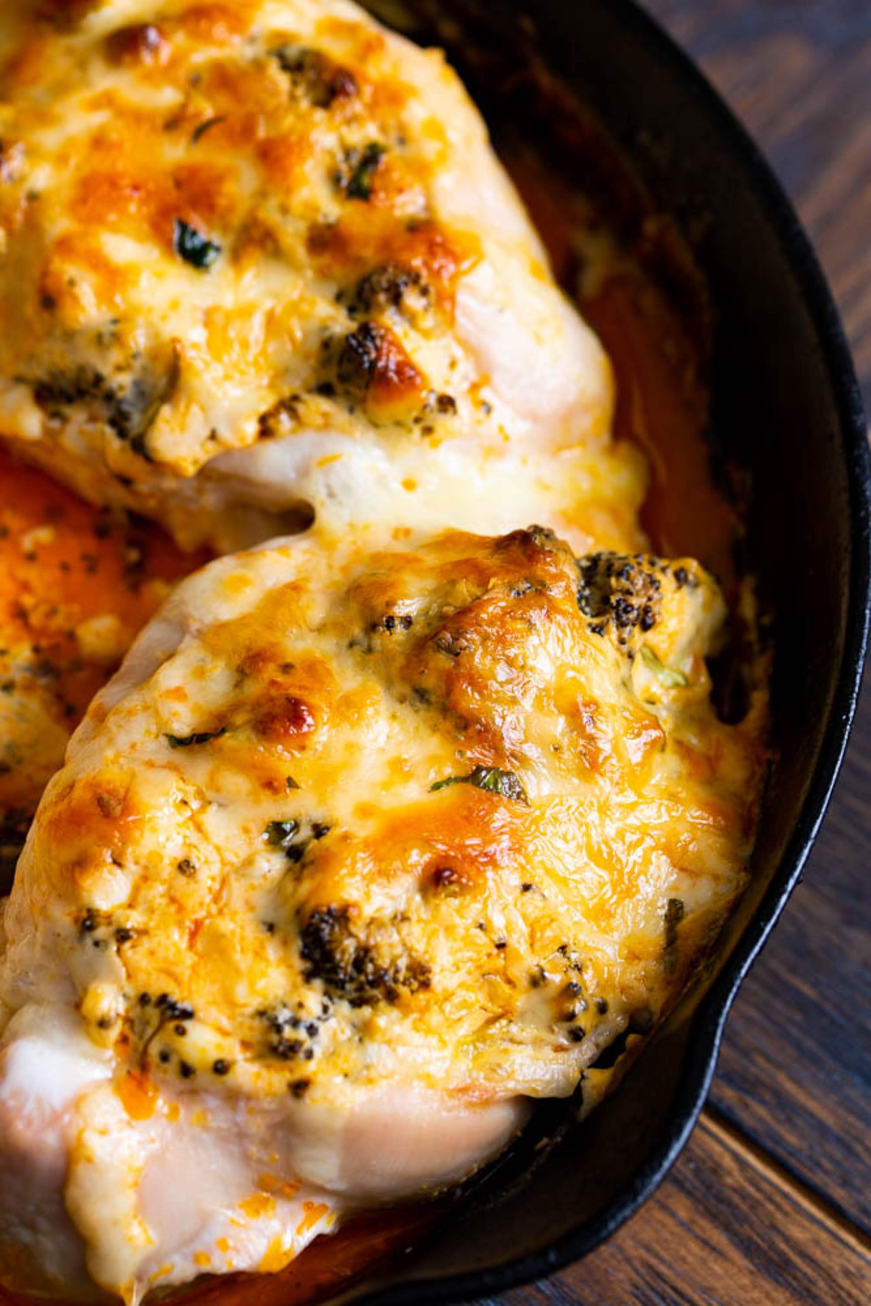 Close-up of two baked chicken breasts in a cast iron skillet, topped with melted cheese and herbs. The dish has a golden-brown crust with visible spices and is sitting on a wooden surface.