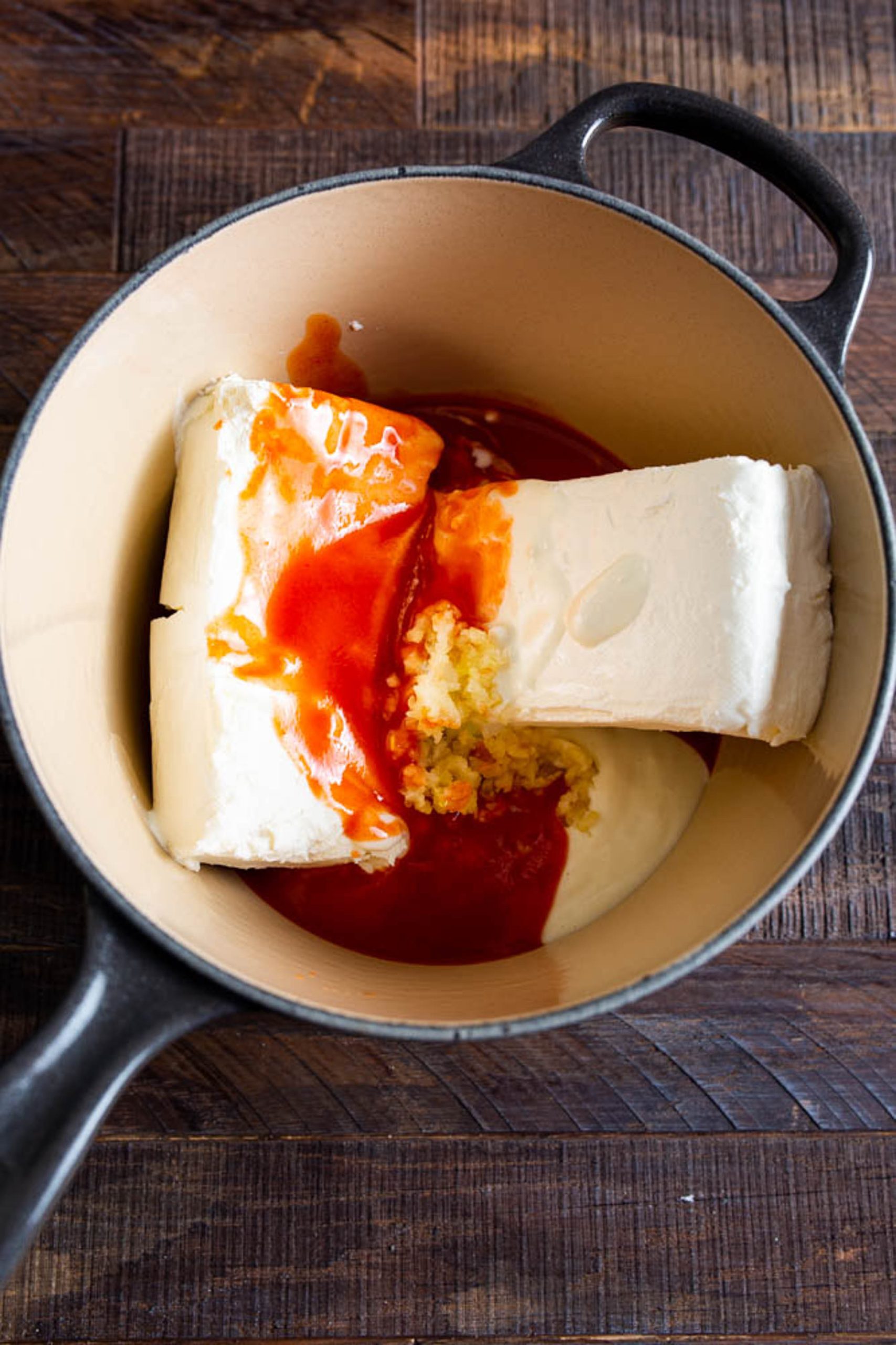 A pot containing blocks of cream cheese, minced garlic, hot sauce, and mayonnaise ingredients on a wooden surface.