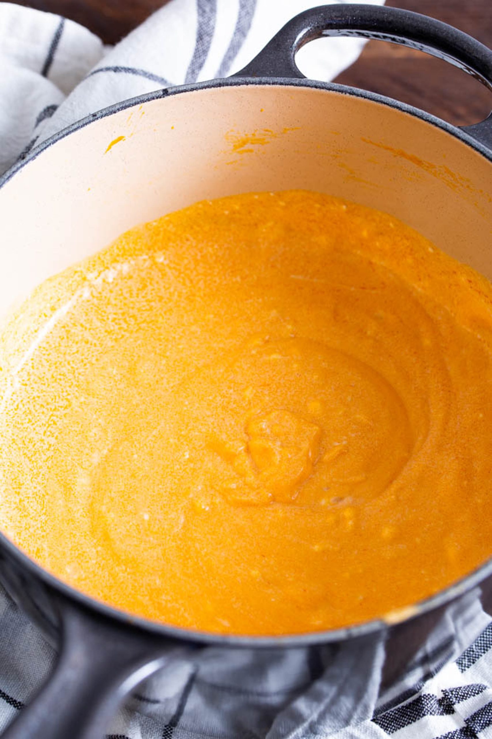 A pot containing a thick, smooth, orange soup or sauce is placed on a striped cloth.