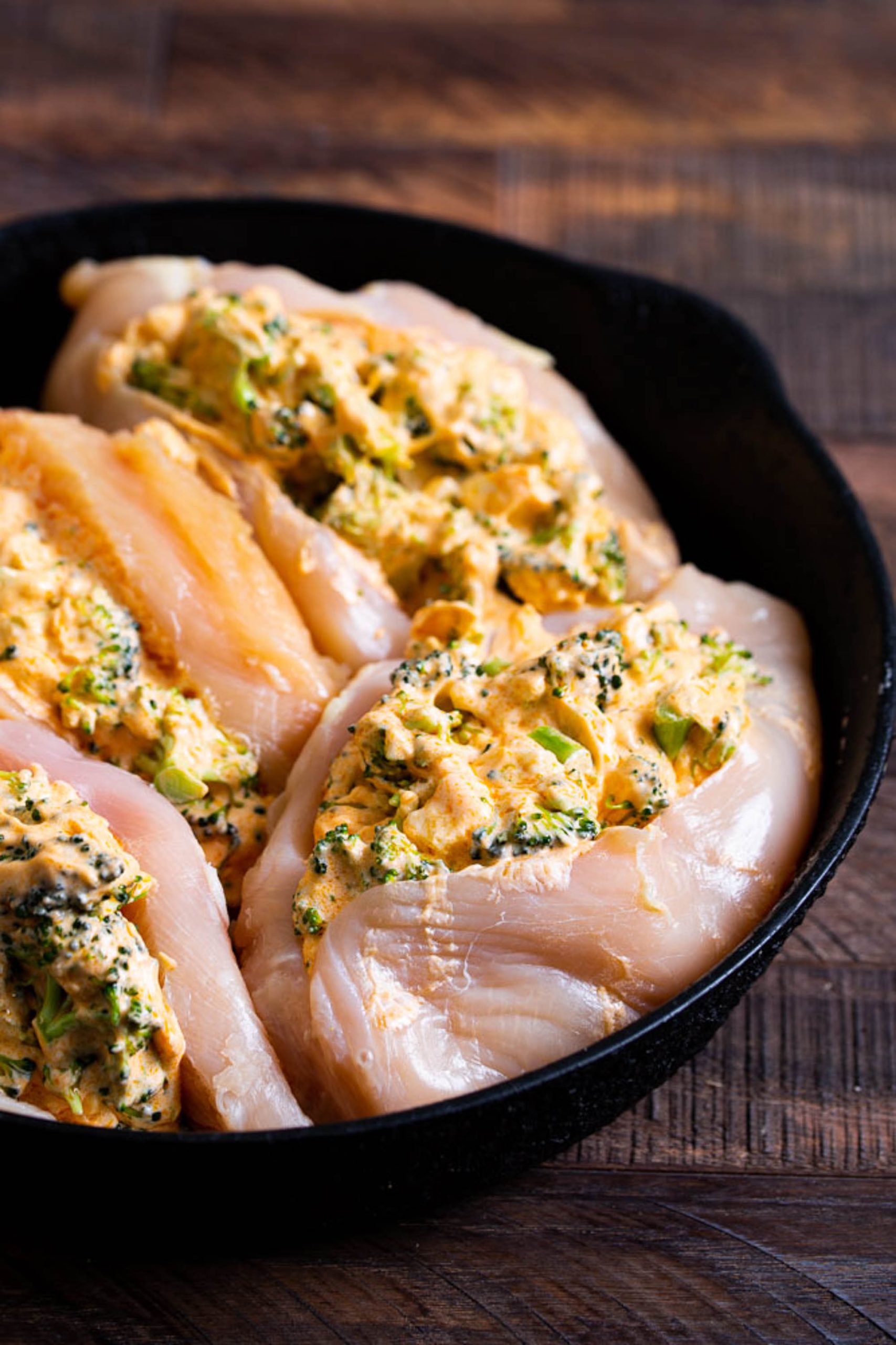 Four raw chicken breasts stuffed with a broccoli and cheese mixture are placed in a black skillet on a wooden surface.