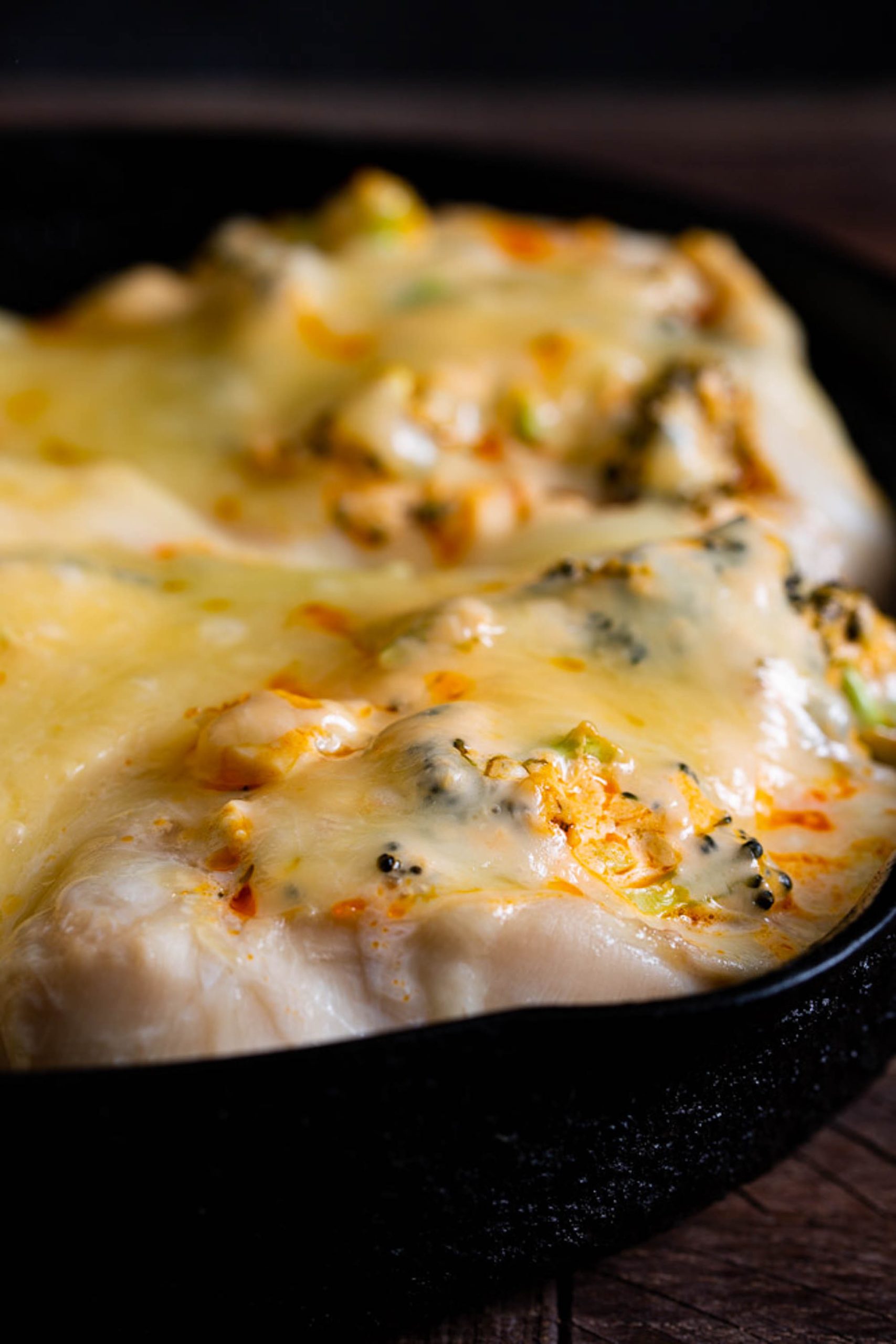 Close-up of cheesy baked dish with visible toppings, including what appears to be broccoli, in a black skillet.