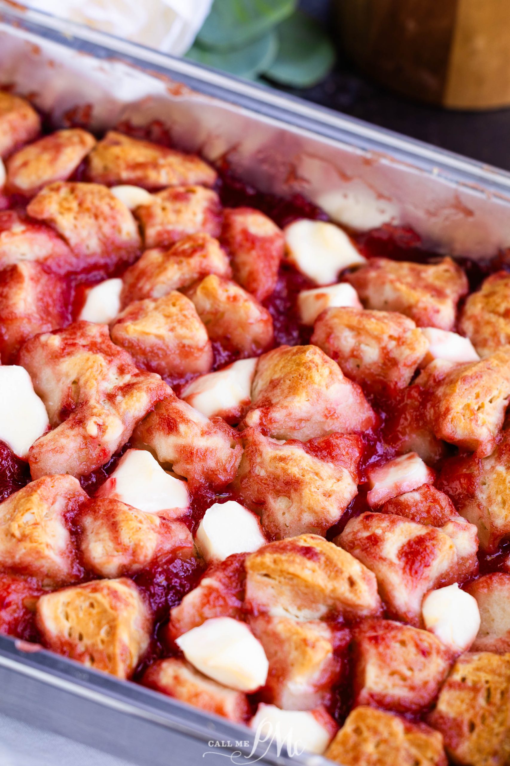 A close-up of a baking dish with a strawberry shortcake casserole, featuring golden-brown biscuit pieces and melted white chocolate over a strawberry sauce.