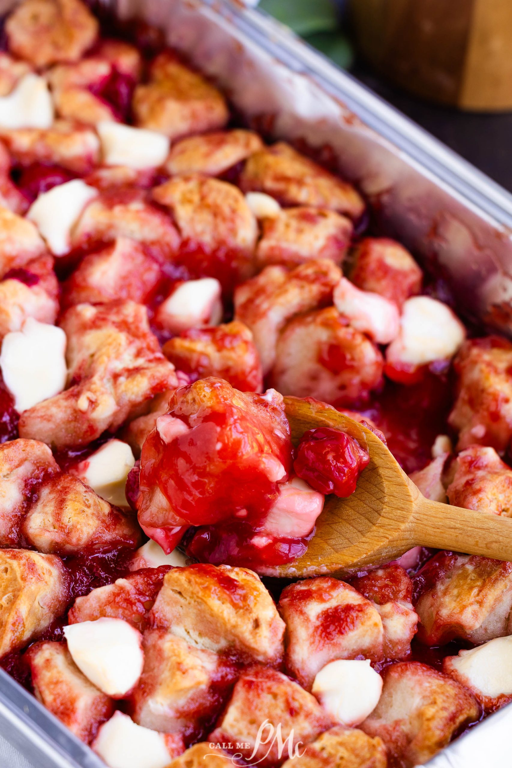 A close-up of a baking dish filled with a bread-based dessert topped with a red fruit sauce and chunks of white cheese, being stirred with a wooden spoon.