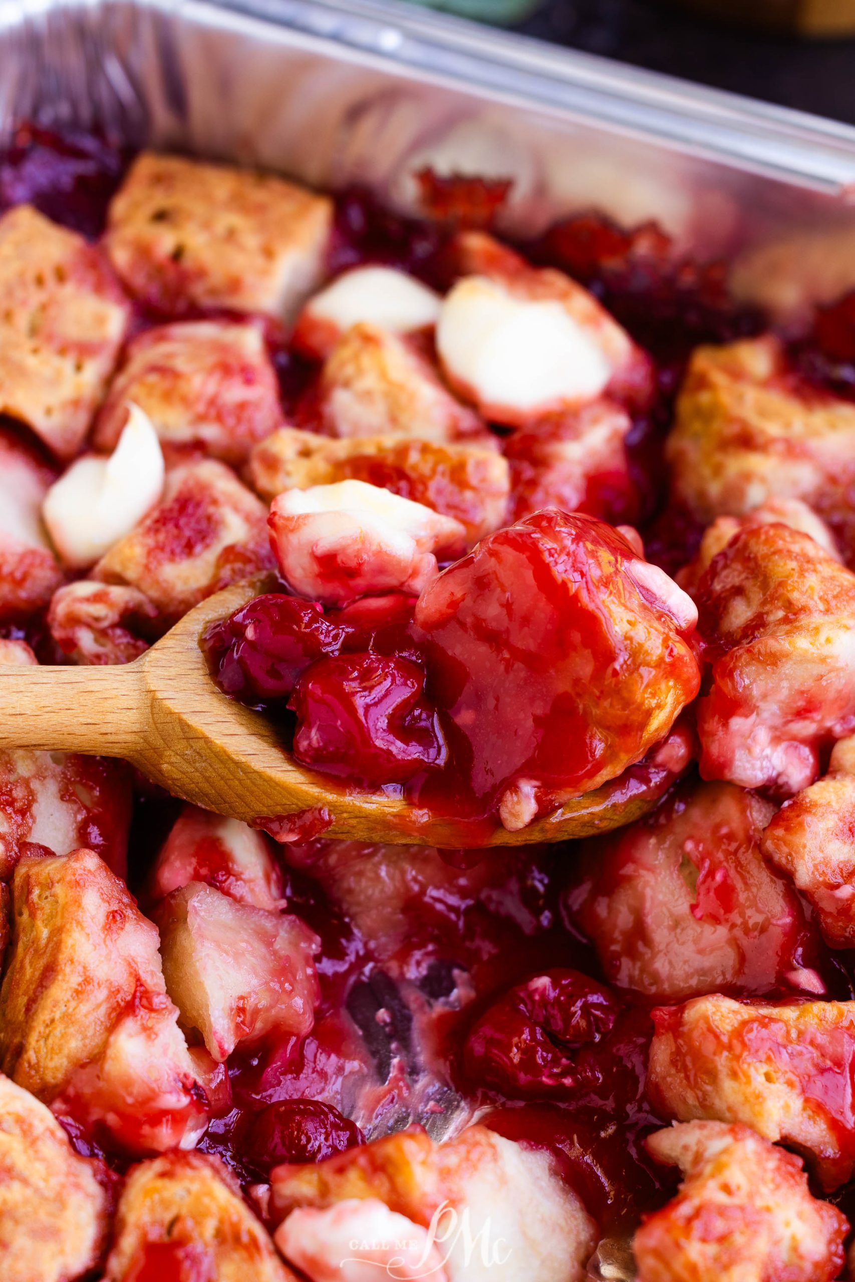 A close-up of a wooden spoon scooping a piece of cherry bread pudding from a pan. The pudding contains visible cherries and custard-soaked bread cubes.
