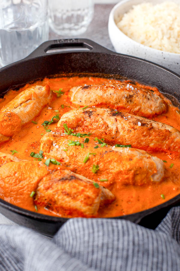 A cast iron skillet filled with cooked chicken breasts in a red tomato-based sauce, garnished with herbs. In the background, a bowl of rice and glasses of water are visible.