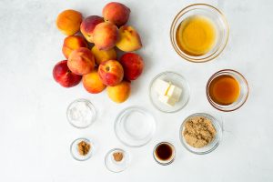 Ingredients for a recipe are laid out on a white surface, including peaches, honey, butter, brown sugar, vanilla extract, spices, and other small ingredients in bowls.