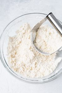 A glass bowl filled with flour and butter mixture being cut with a pastry cutter on a light surface.