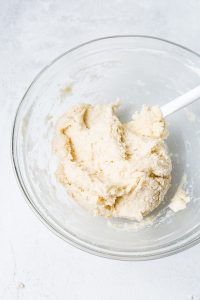 A clear glass bowl with a white spatula contains a mixture of dough on a white surface.