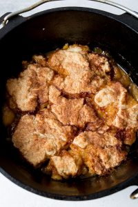 A close-up of a golden brown dessert with a biscuit-like topping and caramelized edges in a dark round baking dish.