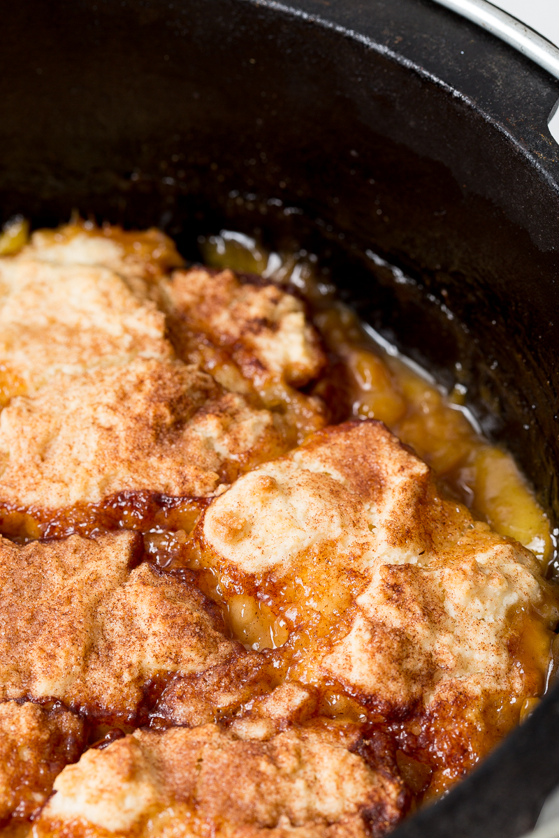 Close-up of a baked cobbler with a golden-brown, sugar-dusted crust in a black baking dish. The filling beneath the crust appears to be fruit-based and slightly bubbling.