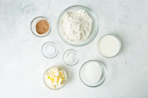 Seven clear glass bowls containing measured baking ingredients including flour, sugar, butter, cinnamon, milk, salt, and baking powder are arranged on a light-colored surface.