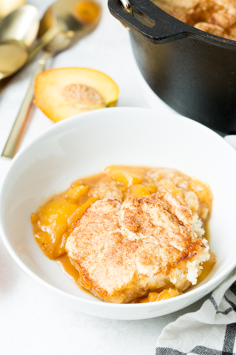 A white bowl filled with peach cobbler topped with cinnamon sugar, placed on a white surface. A slice of peach and a black pot containing more cobbler are seen in the background.