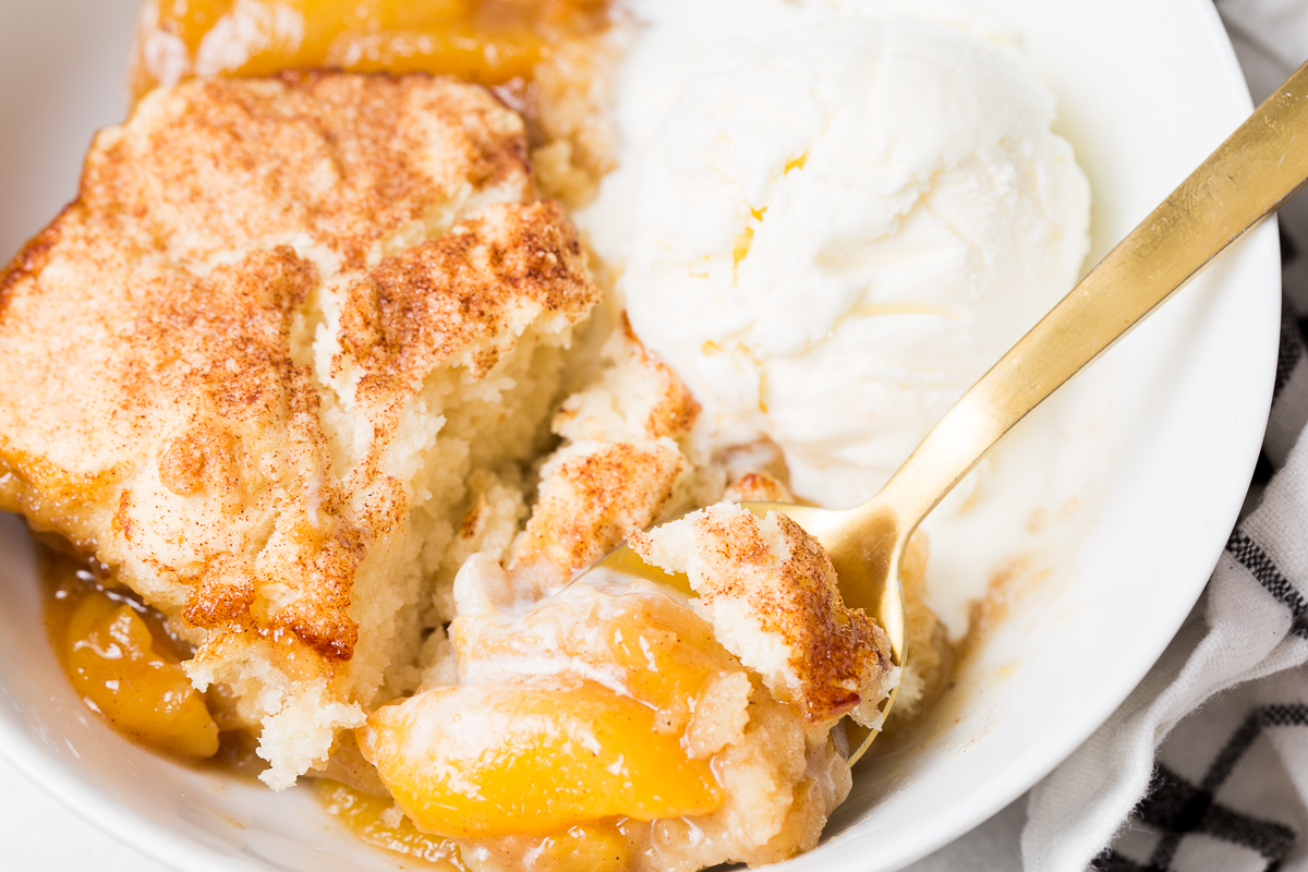 Close-up of a white bowl containing peach cobbler topped with a scoop of vanilla ice cream. A gold spoon is lifting a portion of the cobbler. A checkered cloth is visible in the bottom right.