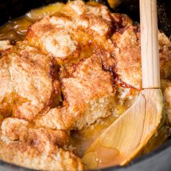 A close-up of a peach cobbler in a cast iron skillet. The golden-brown crust is partially scooped to reveal the juicy peach filling underneath. A wooden spoon rests at the edge.