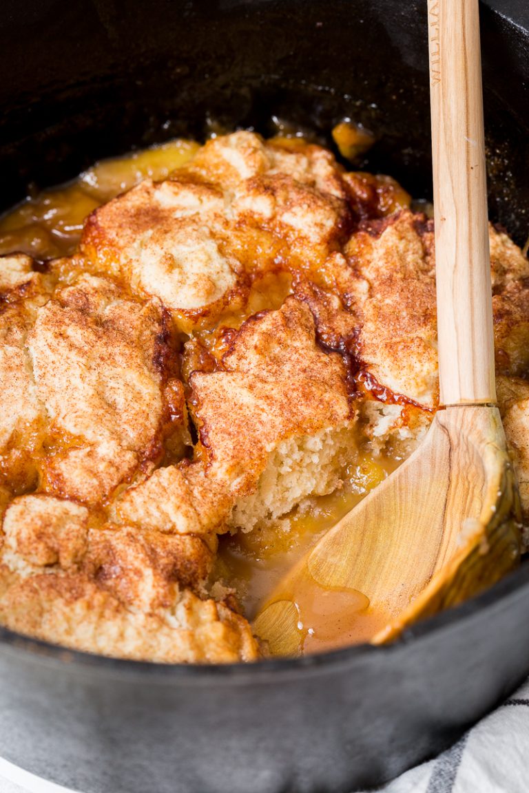 A close-up of a peach cobbler in a cast iron skillet. The golden-brown crust is partially scooped to reveal the juicy peach filling underneath. A wooden spoon rests at the edge.