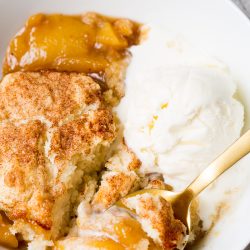 A bowl contains peach cobbler topped with a sprinkle of cinnamon and sugar, served alongside a scoop of vanilla ice cream. A gold spoon rests inside the bowl.