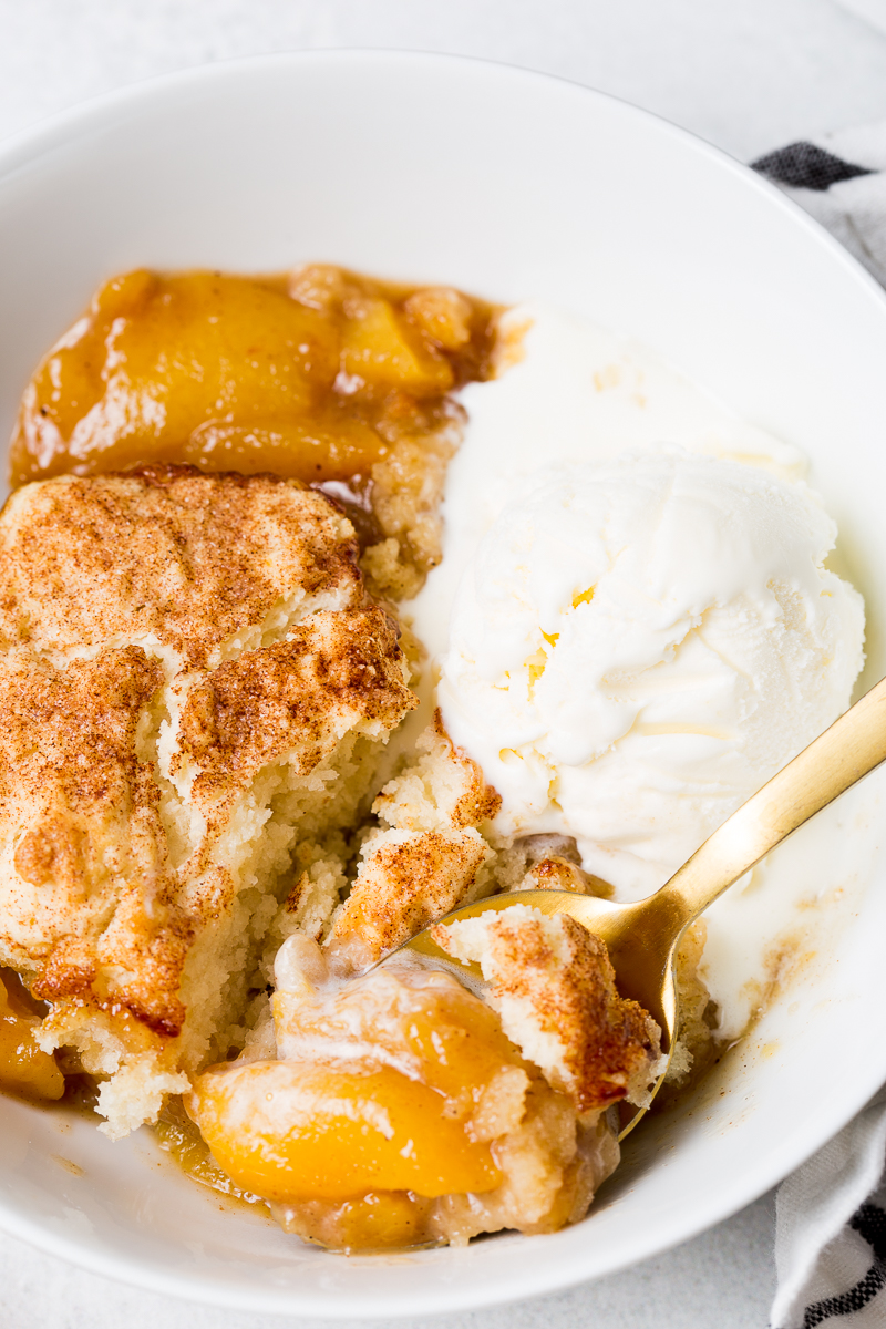 A bowl contains peach cobbler topped with a sprinkle of cinnamon and sugar, served alongside a scoop of vanilla ice cream. A gold spoon rests inside the bowl.