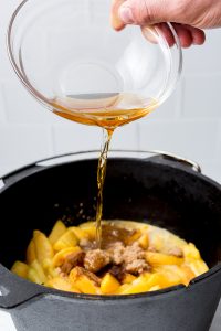 A hand pours liquid from a glass bowl into a pot filled with sliced peaches, brown sugar, and spices.