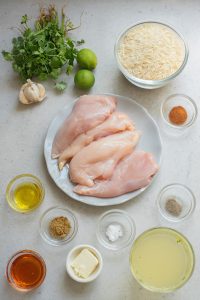 Ingredients for a dish include raw chicken breasts, bowls of rice, garlic, limes, cilantro, spices, olive oil, broth, butter, honey, salt, and pepper arranged on a countertop.