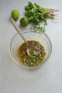 A clear bowl containing a mixed herb and spice marinade with a spoon, placed on a light countertop. Nearby are two whole limes and a small bunch of cilantro with roots attached.