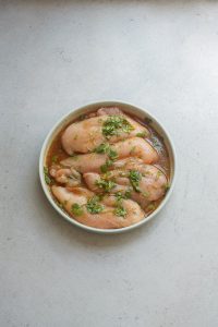 A white bowl filled with raw chicken breasts marinating in a seasoned liquid with green herbs on a light grey countertop.