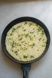 Cooked rice with herbs in a black frying pan on a light gray countertop.