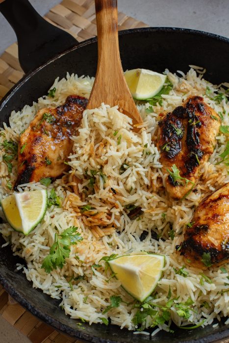 A skillet filled with cooked rice, grilled chicken pieces, cilantro, and lime wedges. A wooden spoon is seen mixing the ingredients.