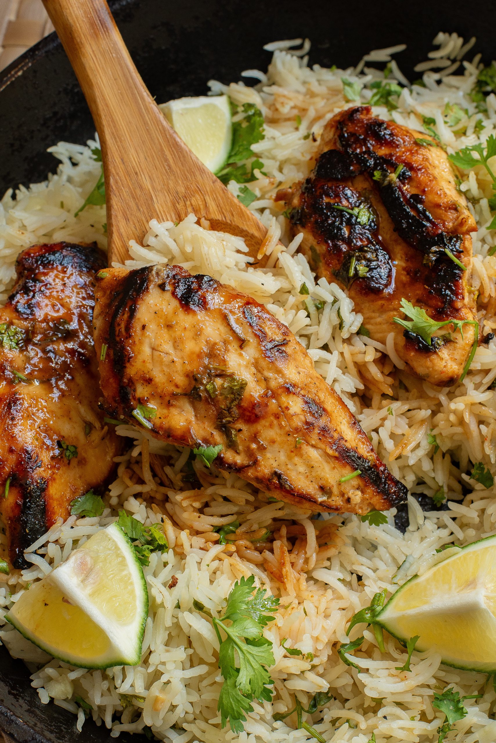 A close-up of grilled chicken breasts on a bed of seasoned rice, garnished with cilantro and lime wedges, being stirred with a wooden spoon.