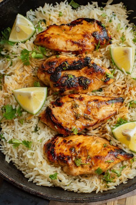 A pan filled with seasoned grilled chicken breasts on a bed of white rice, garnished with lime wedges and fresh herbs.