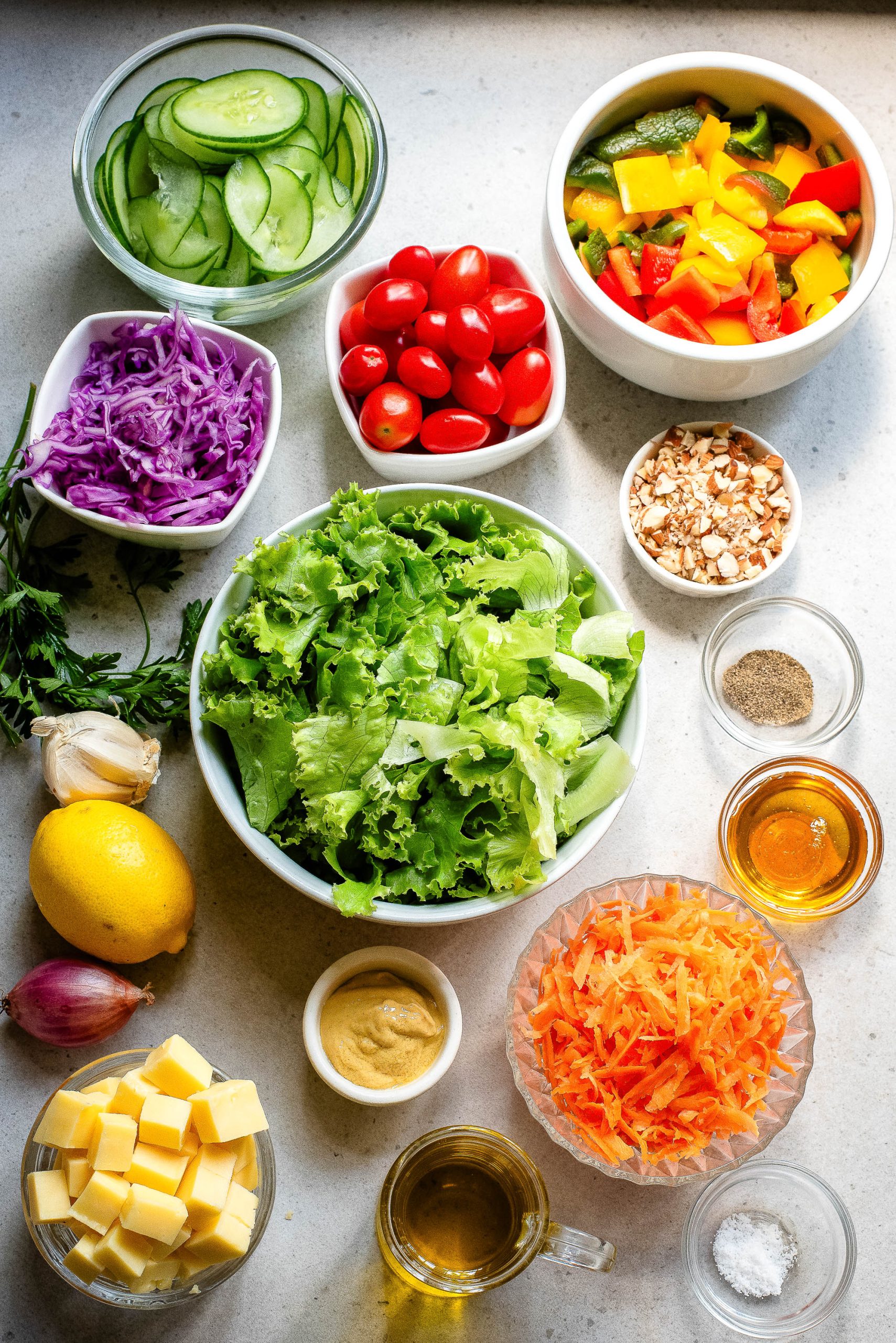 An assortment of fresh ingredients displayed on a kitchen counter, including lettuce, cherry tomatoes, sliced cucumbers, shredded carrots, diced bell peppers, red cabbage, garlic, lemon, and various seasonings.