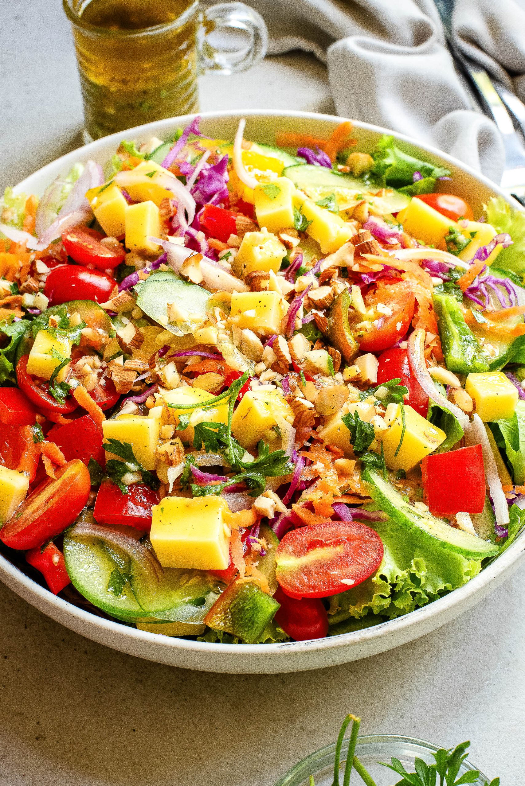 A colorful mixed vegetable salad in a white bowl, featuring diced mango, cherry tomatoes, bell peppers, cucumber slices, red cabbage, and greens, with a jug of dressing in the background.