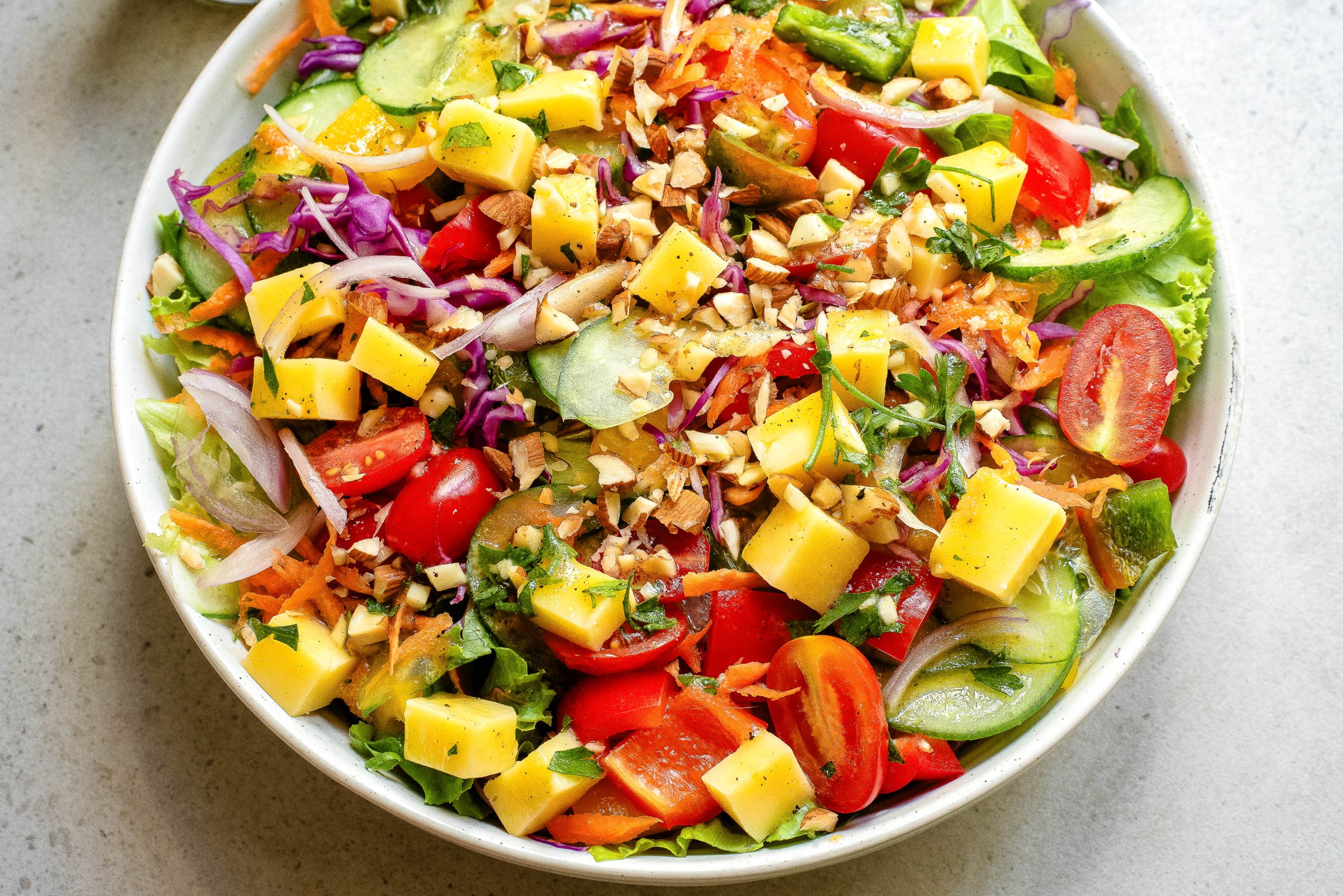 A colorful salad in a white bowl featuring mixed greens, cherry tomatoes, cucumber slices, diced mango, shredded carrots, purple cabbage, chopped nuts, and herbs.