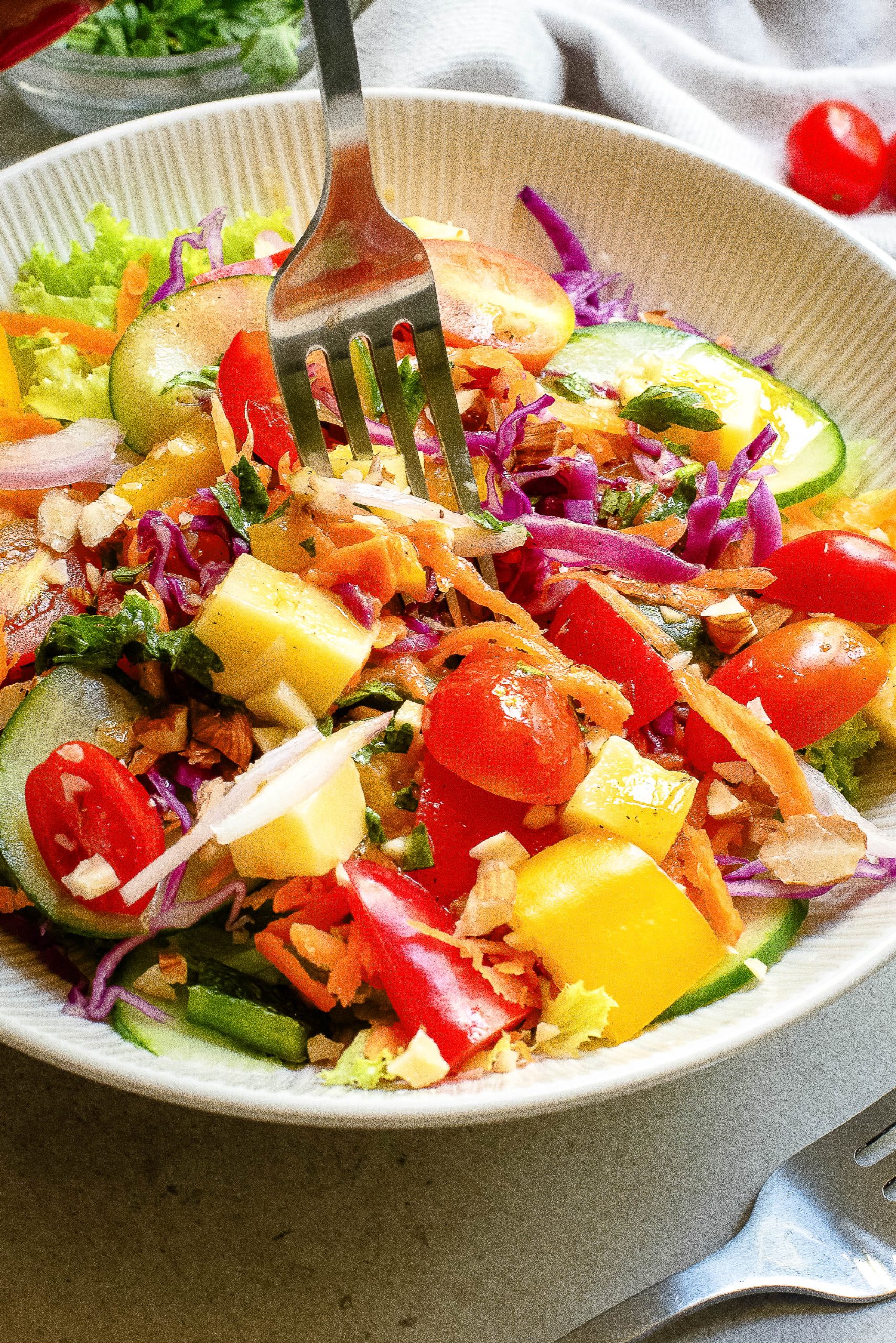 A colorful salad with cherry tomatoes, cucumber slices, red cabbage, yellow bell peppers, shredded carrots, and chopped nuts is being mixed with a fork in a white bowl.