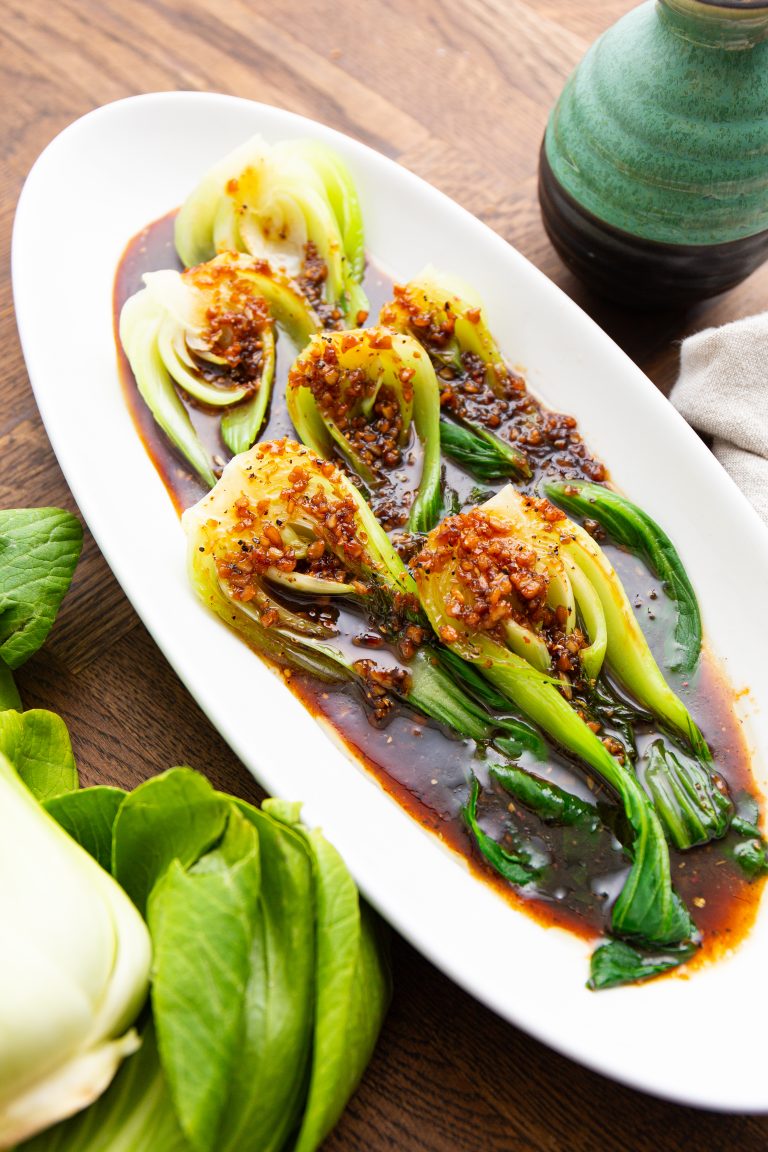 A plate of sautéed bok choy drizzled with a spicy garlic sauce, garnished with red pepper flakes, and accompanied by a green ceramic condiment container.