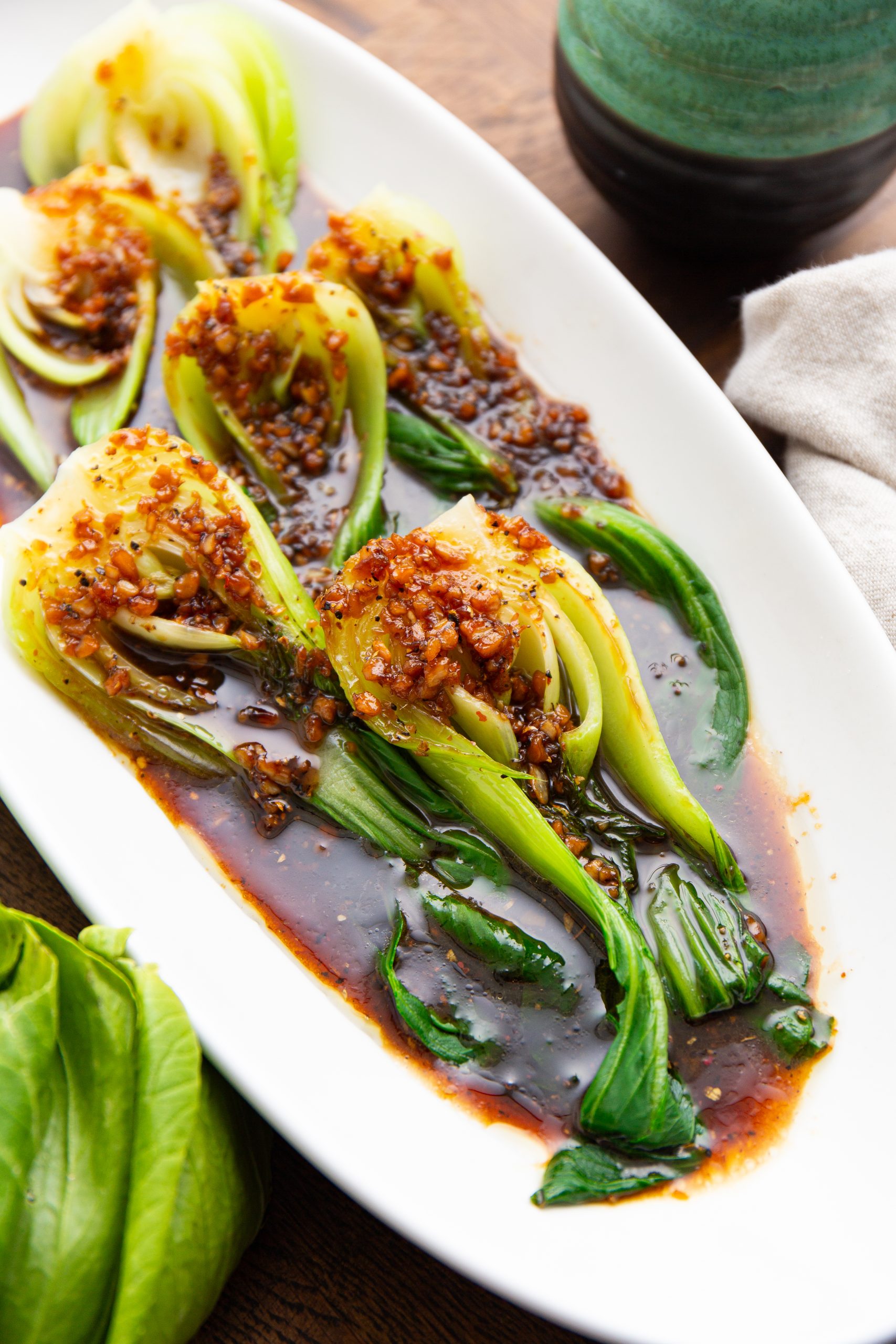 A plate of sautéed bok choy drizzled with a spicy garlic sauce, garnished with red pepper flakes, and accompanied by a green ceramic condiment container.