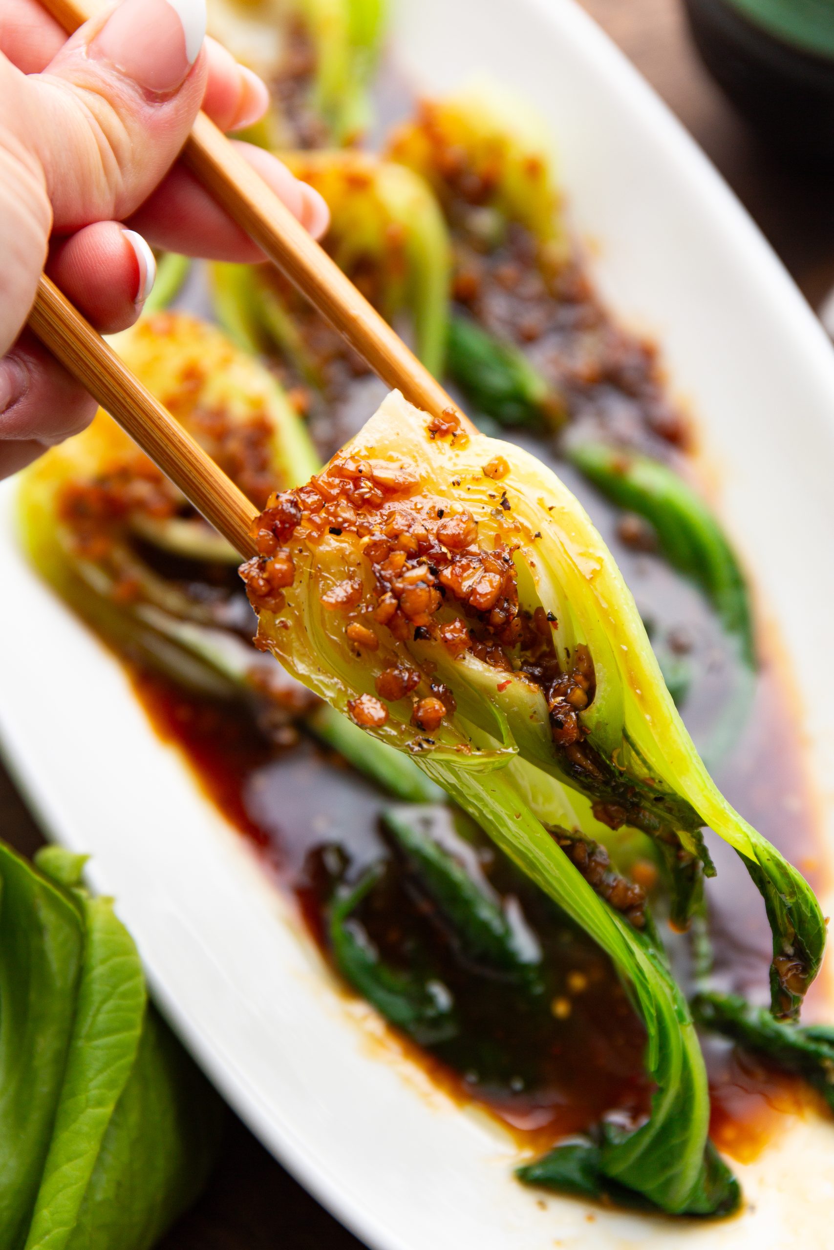 A hand holding chopsticks lifts a piece of steamed vegetable from a white plate garnished with dark soy-based sauce.