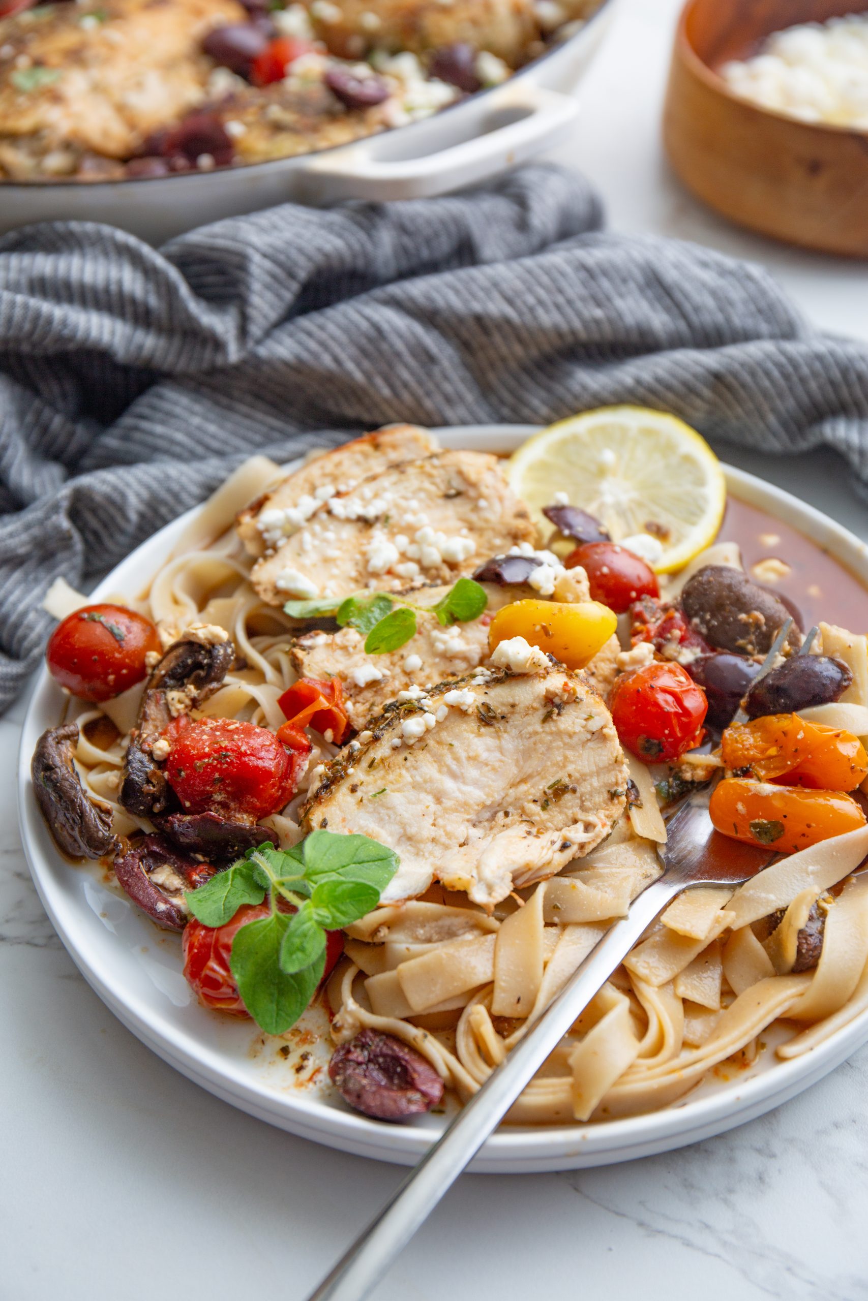 A pan of cooked chicken breasts garnished with olives, cherry tomatoes, feta cheese crumbles, and herbs.