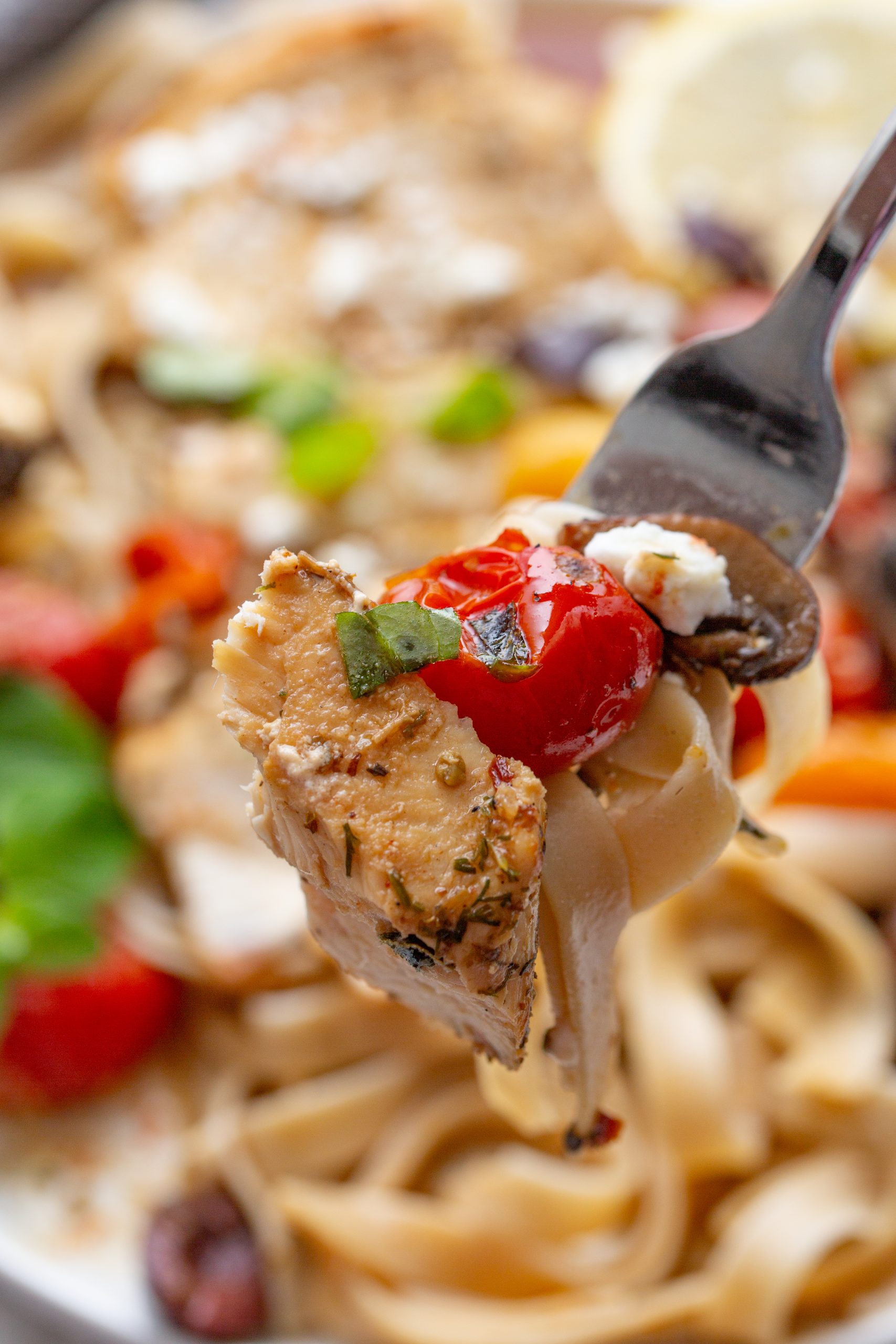 A fork holding a bite of pasta topped with a piece of seasoned chicken, a cherry tomato, and garnished with herbs. Blurred background shows more pasta and vegetables.