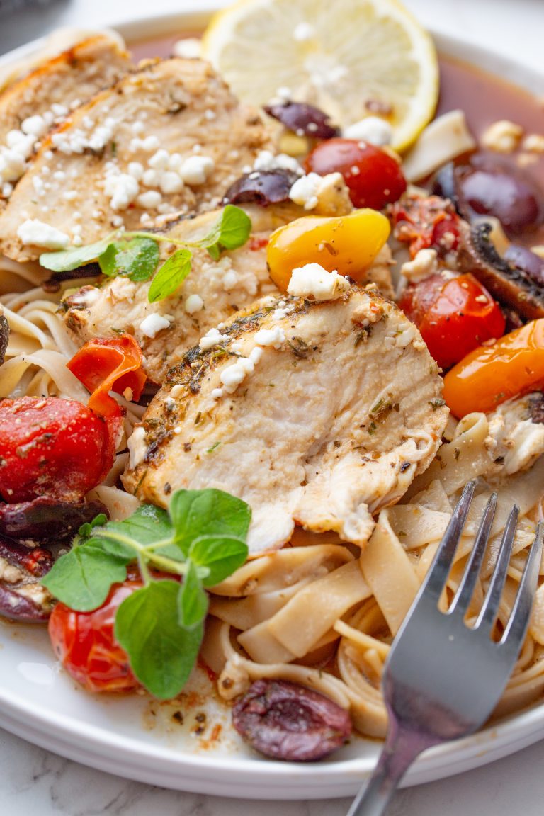 A plate of pasta topped with sliced grilled chicken, cherry tomatoes, Kalamata olives, feta cheese, fresh herbs, and a lemon slice. A fork rests on the plate.