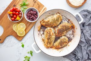 A white pan containing four seasoned and roasted chicken breasts, with small bowls of cherry tomatoes, lemon slices, and olives on a wooden cutting board nearby. A striped cloth is on the right side.