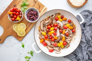 Sautéed mushrooms and cherry tomatoes in a white pan on a marble surface, accompanied by sliced lemons, olives, and herbs on a wooden board. Bowl of cheese and a striped cloth nearby.