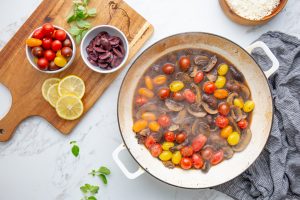 A pan of cherry tomatoes and mushrooms cooking, placed on a marble countertop with small bowls of sliced olives, cherry tomatoes, lemon slices, and fresh herbs on a wooden cutting board nearby.