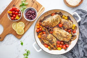 A cooked dish of seasoned chicken breasts with cherry tomatoes and mushrooms in a pan, next to a cutting board with bowls of cherry tomatoes and olives, lemon slices, and herbs on a marble surface.