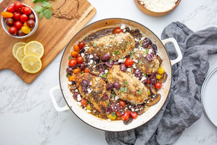 A skillet contains baked chicken breasts with cherry tomatoes, olives, mushrooms, and crumbled cheese on a marble surface with lemon slices and a wooden board.