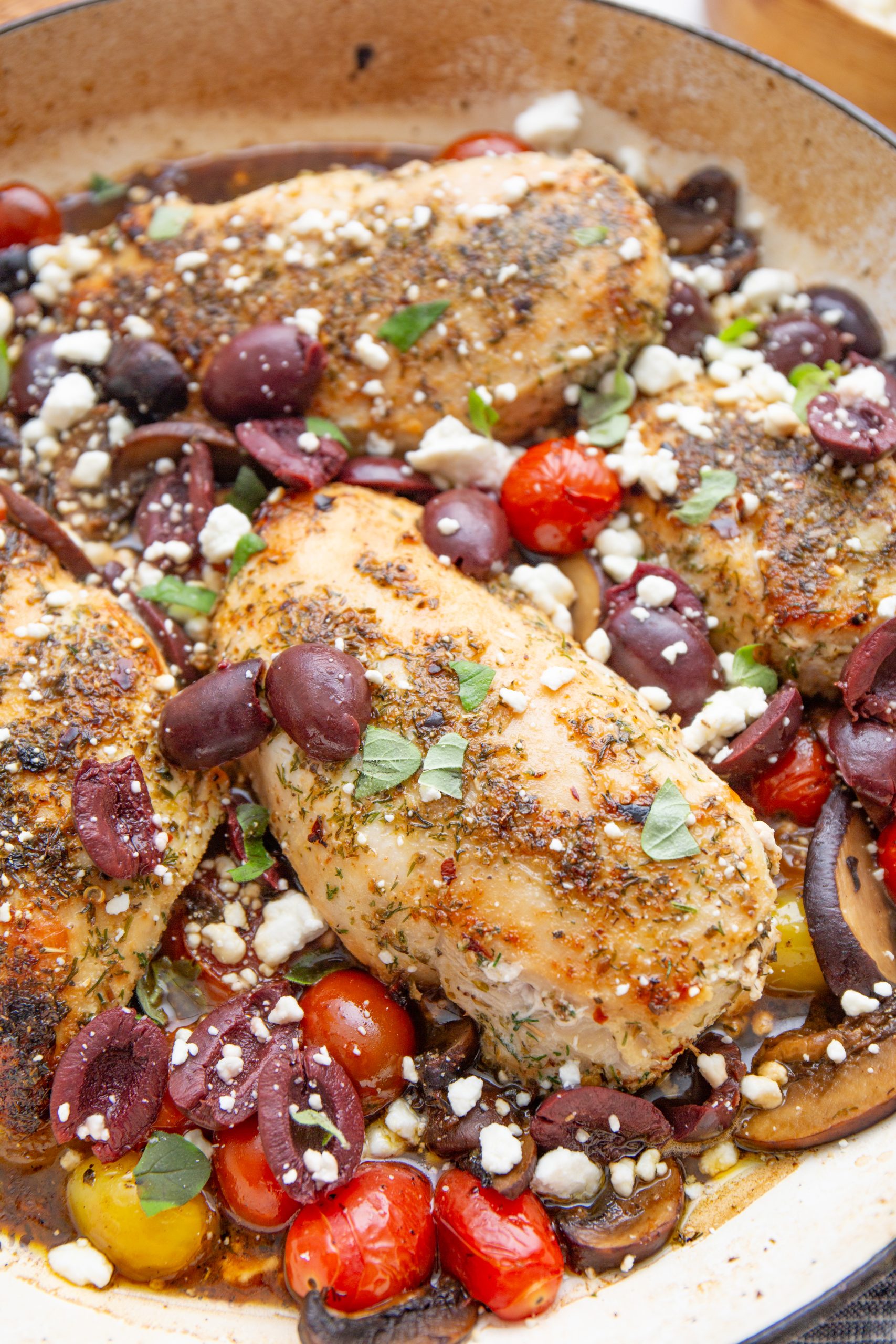 A pan of cooked chicken breasts garnished with olives, cherry tomatoes, feta cheese crumbles, and herbs.