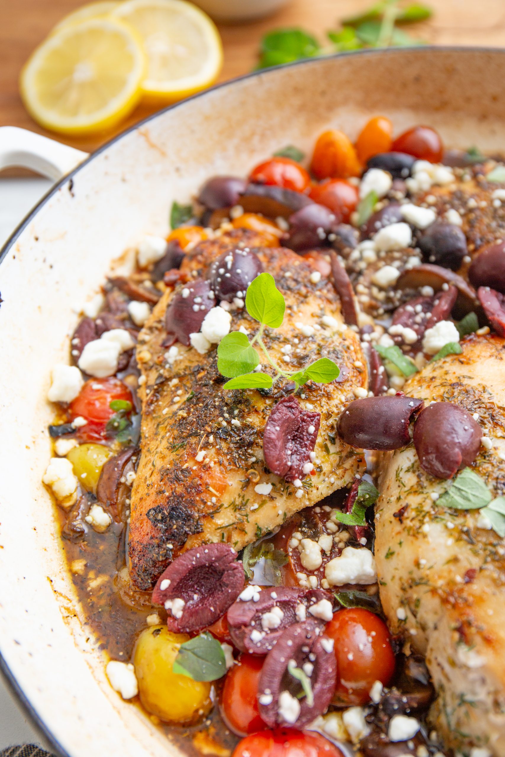 A pan of cooked chicken breasts garnished with olives, cherry tomatoes, feta cheese crumbles, and herbs.